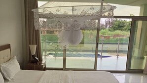 Ein Schlafzimmer mit einem großen Bett, einem Nachttisch, einer Lampe und Blick auf einen Swimmingpool durch eine Glastür.
