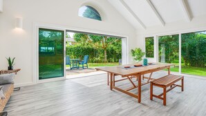 Ruime kamer met een houten tafel en stoelen, grote schuifdeuren die uitkomen op een tuin, en een hoog plafond met zichtbare balken.