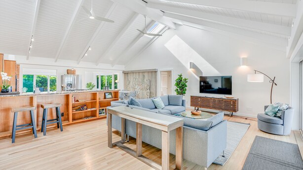 Een ruime woonkamer met een hoge plafond, houten vloeren, een zithoek met een bank en salontafel, een keuken met houten kastjes, en een groot raam dat natuurlijk licht binnenlaat.