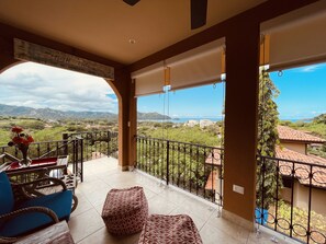 Un balcón con vistas a un paisaje montañoso, piso de baldosas y una zona de estar con cojines y una mesa.