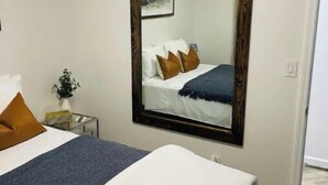 Un dormitorio con un espejo grande, una cama con ropa de cama blanca, un plaid azul y una mesita de noche pequeña con una planta.