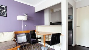 Un espacio habitable compacto con un sofá, dos sillones, una mesita, una lámpara de pie y una kitchenette al fondo.