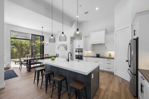A modern kitchen with a large island, bar stools, pendant lighting, and a view of the outdoors. - 2023 Captiva Home | Steps to The Beach/Shops/Dining (Marco Island)