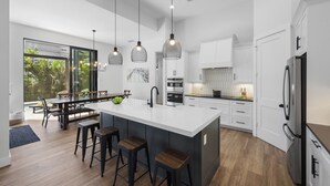 A modern kitchen with a large island, bar stools, pendant lighting, and a view of the outdoors. - 2023 Captiva Home | Steps to The Beach/Shops/Dining (Marco Island)