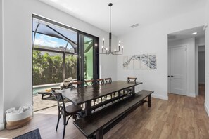A modern dining area with a large wooden table, benches, and chairs. It features a large window with a view of greenery and a pool outside. - 2023 Captiva Home | Steps to The Beach/Shops/Dining (Marco Island)
