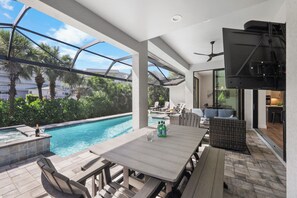 An outdoor pool area with a wooden table and chairs, a ceiling fan, and a view of the pool and surrounding greenery. - 2023 Captiva Home | Steps to The Beach/Shops/Dining (Marco Island)