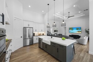 A modern kitchen with stainless steel appliances, a large island, pendant lights, and a dining area with a sofa and TV. - 2023 Captiva Home | Steps to The Beach/Shops/Dining (Marco Island)