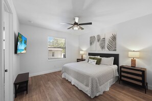 A bedroom with a bed, a ceiling fan, a nightstand, a lamp, a TV, and a window with blinds.