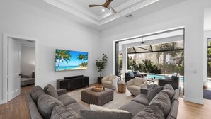 A spacious living room with a large gray sectional sofa, a flat-screen TV mounted on the wall, and a view of a pool through large windows. - 2023 Captiva Home | Steps to The Beach/Shops/Dining (Marco Island)