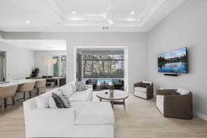 A modern living room with a white sofa, a coffee table, and a TV on the wall. There is a dining area with wooden chairs and a table, and a view of a pool and greenery outside.