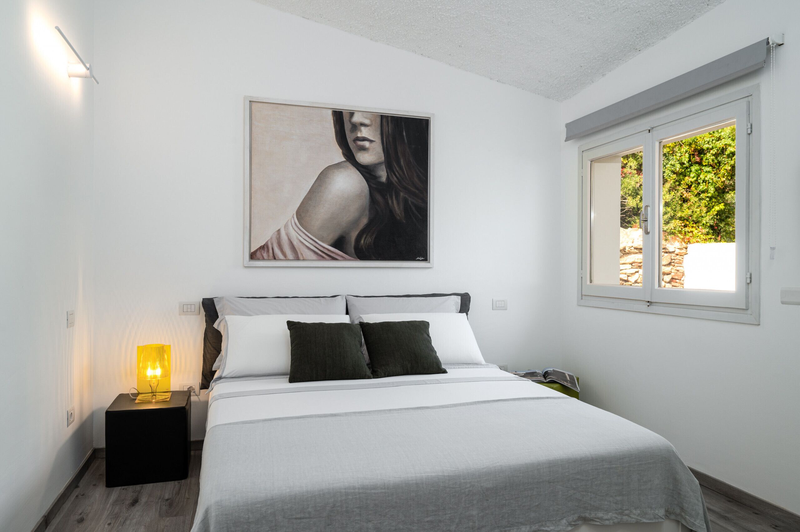A modern bedroom with a bed, a nightstand with a lamp, a painting on the wall, and a window with a view of greenery.