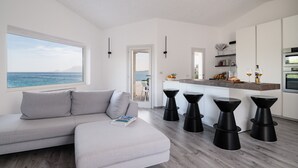 A modern living room with a large window overlooking the sea, a sofa with a magazine, and a kitchen area with bar stools and a countertop.