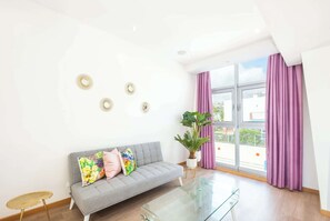 Een moderne woonkamer met een grijze bank, kleurrijke kussens, een glazen salontafel en een groot raam met paarse gordijnen.
