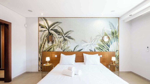 Habitació d'hotel amb un llit gran, un panell de fusta al capçal, dues taules de nit amb làmpades i un mural amb temàtica tropical a la paret.