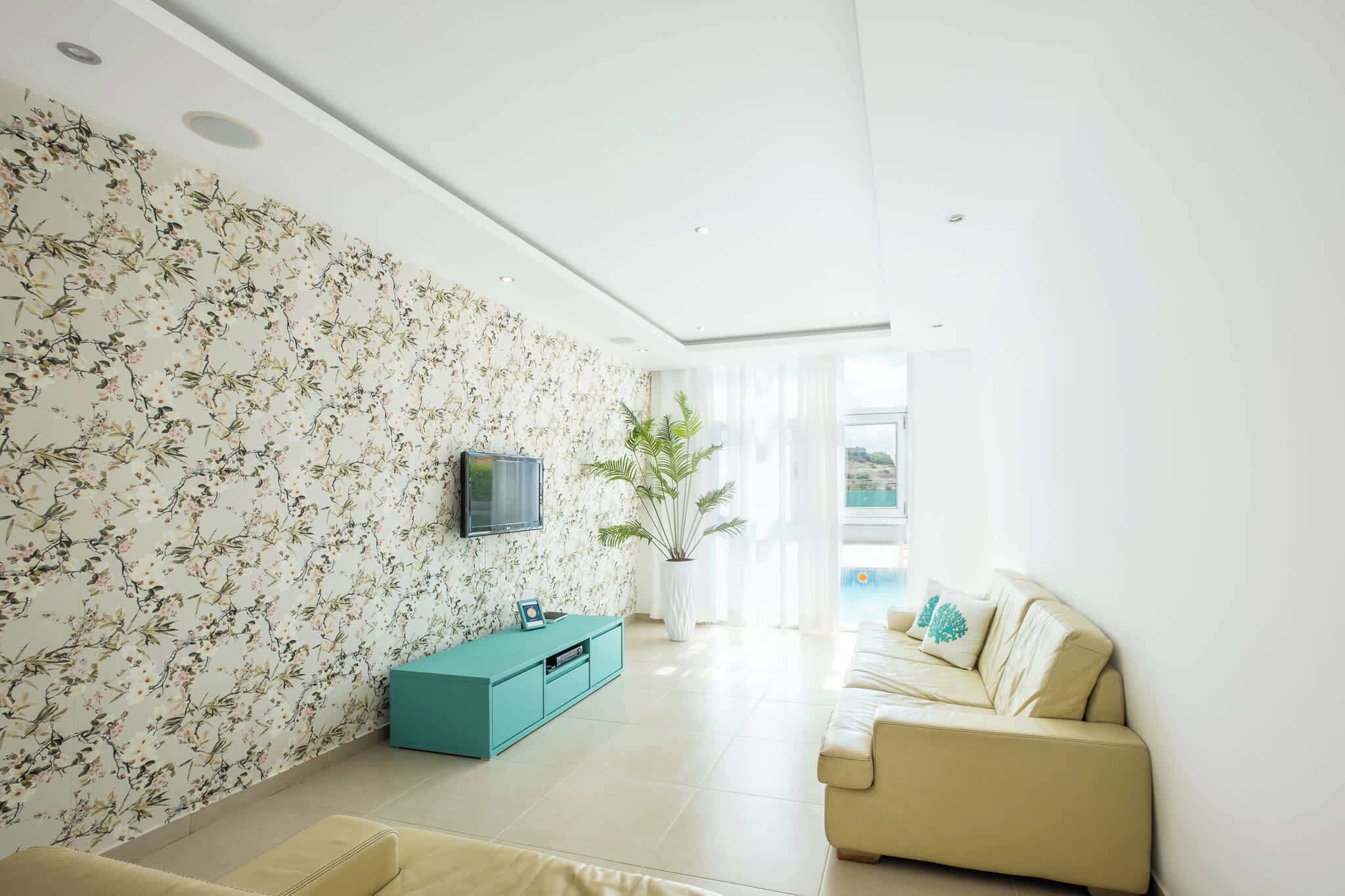 A modern living room with a floral wallpaper, a teal sideboard, a beige sofa, and a large window providing natural light.