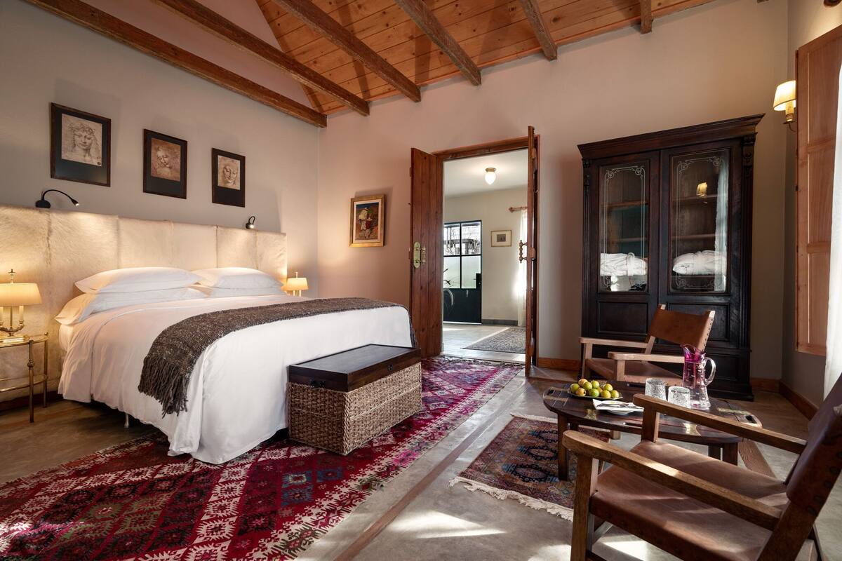 A bedroom with a large bed, a wooden headboard, a bedside table with a lamp, a small bench, a chair, a cabinet, and a red patterned rug.