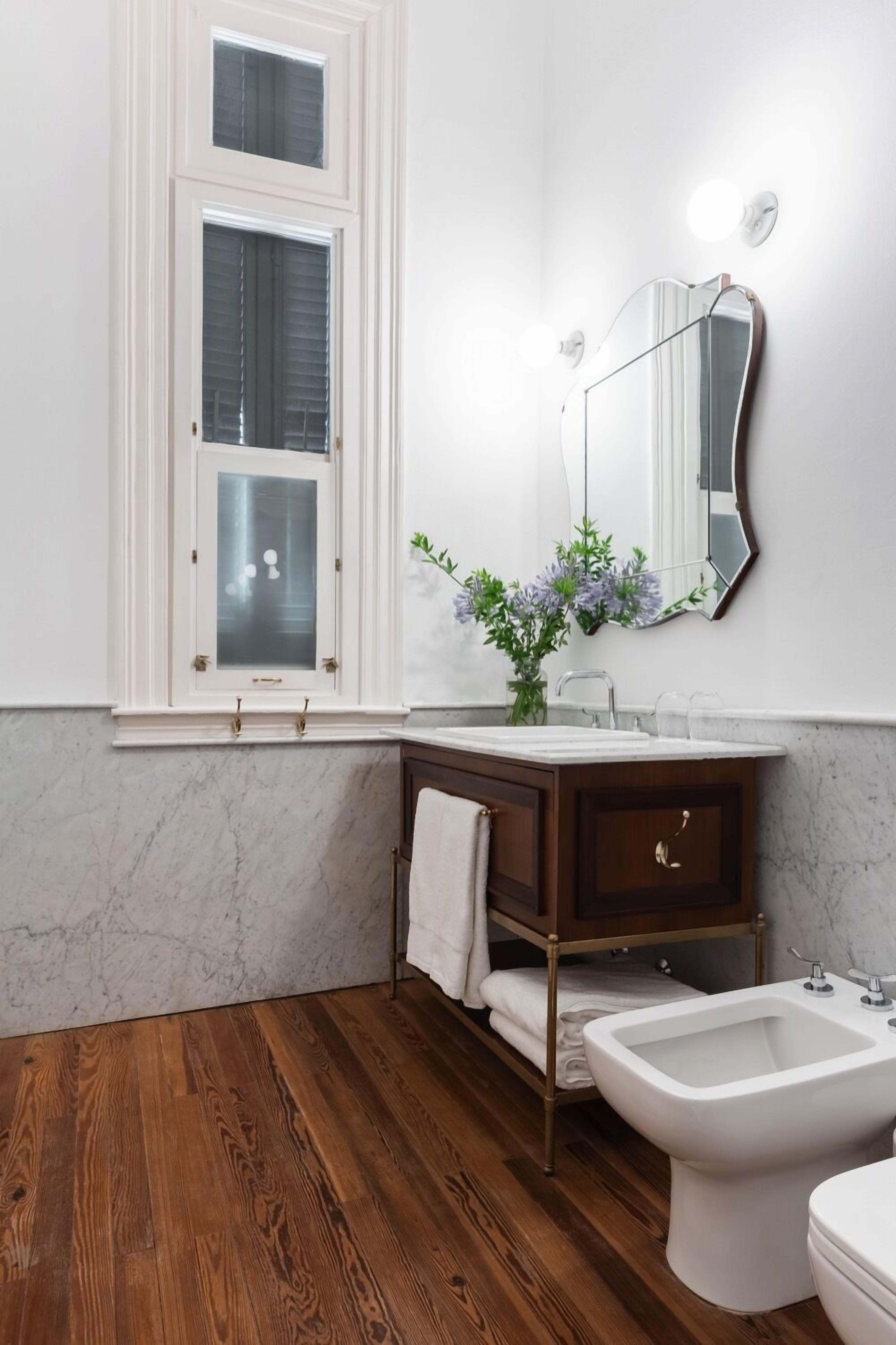 A bathroom with a wooden floor, white marble walls, a white vanity with a mirror, and a white bidet.