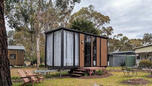 A small wooden cabin with a sliding door, a picnic table, and a bench in a grassy area with trees. - "Timboon" Tiny House 10 - Escape @ Grampians' Edge (Dadswells Bridge)
