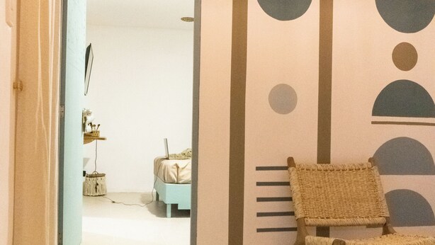 A hallway with a wicker chair, a bed with a laptop, and a wall with geometric patterns. - Chaim, Happiness to be where you should be. (Campeche)