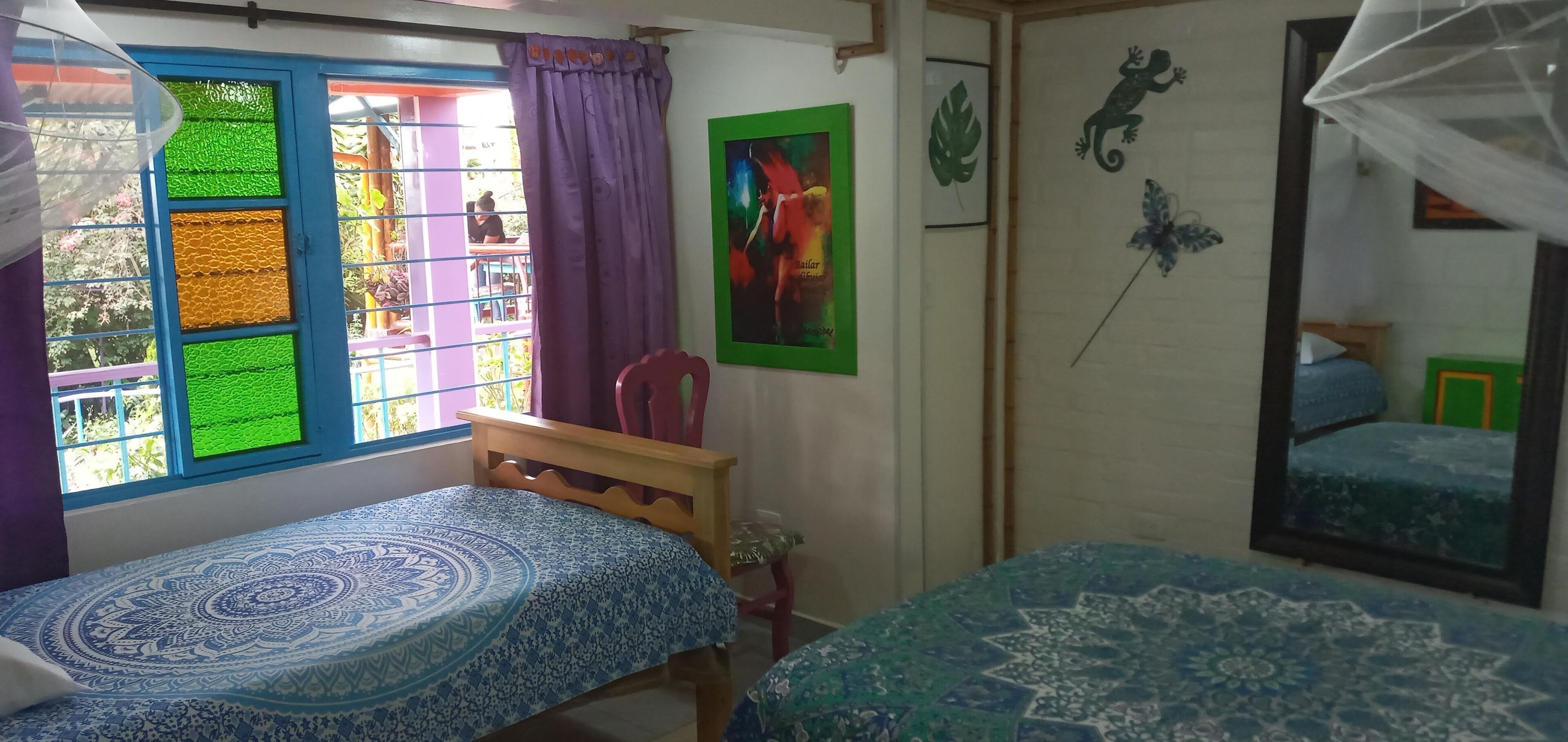 A room with two beds, a window with colorful panes, a chair, and a mirror.