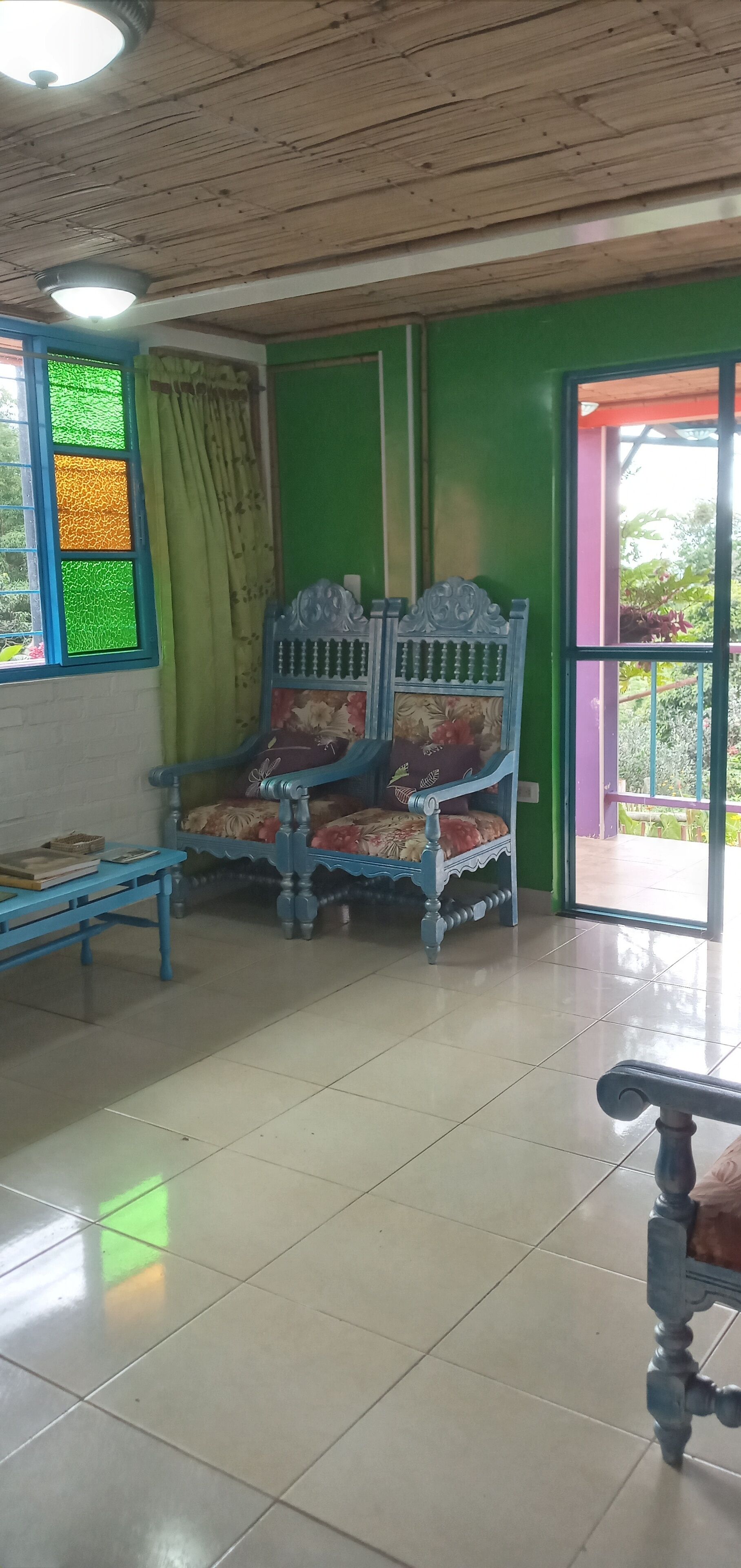 A room with a wooden ceiling, a green wall, a blue bed with floral bedding, a blue table with books, and a window with green and orange panes.