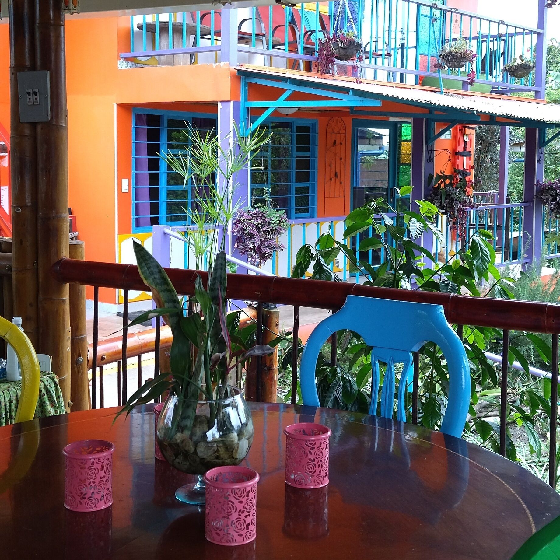 A table with a vase and pink cups, a blue chair, and a balcony with colorful buildings in the background.