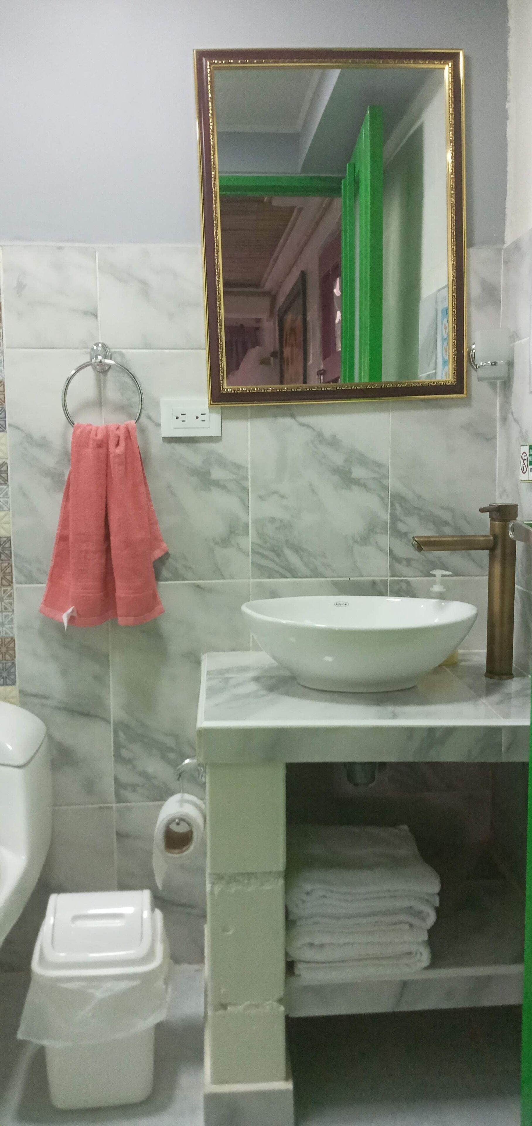 Un baño con pared de azulejos de mármol, un lavamanos blanco con grifo de latón, una toalla rosa colgada en un aro metálico y un espejo con marco dorado.
