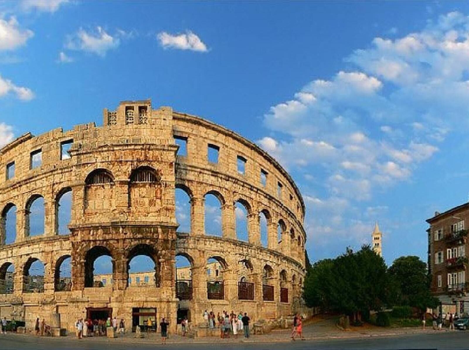 Un amphithéâtre en pierre d’époque, doté de nombreuses arcades et d’une scène centrale, entouré de touristes et de bâtiments modernes.
