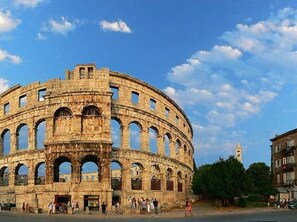 Un amphithéâtre en pierre d’époque, doté de nombreuses arcades et d’une scène centrale, entouré de touristes et de bâtiments modernes.