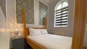 A bedroom with a wooden bed, a nightstand, a lamp, and a window with blinds. - Baylis Park Hotel (Slough)
