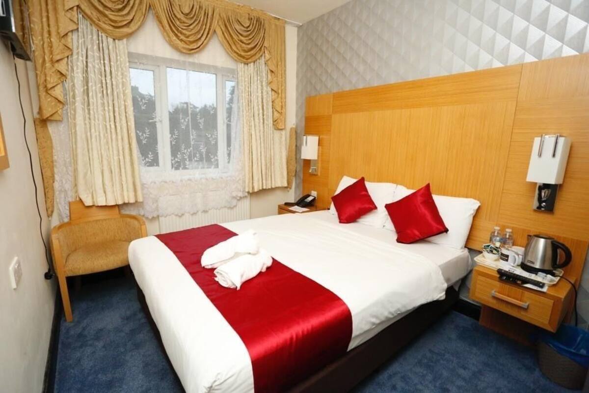 Une chambre d’hôtel avec un grand lit, une literie rouge et blanche, une tête de lit en bois, une table de chevet avec une bouilloire et une chaise.