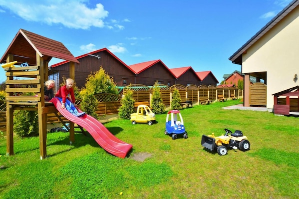 A playground with a red slide and climbing structure, surrounded by wooden playhouses and toy vehicles.