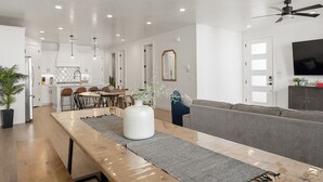 Une cuisine moderne avec des meubles blancs, un coin repas doté d’une table et de chaises en bois, et un canapé gris dans le salon.