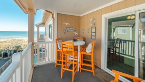 Een balkon met een tafel en stoelen, uitzicht op het strand, en een houten huis op de achtergrond.