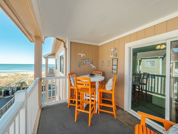 A balcony with a table and chairs, a view of the beach, and a wooden house in the background.