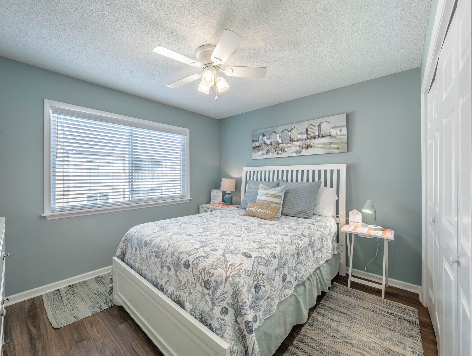 A bedroom with a bed, a ceiling fan, a window with blinds, a nightstand, and a painting on the wall.