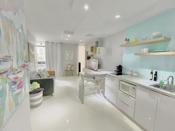A modern kitchen with white cabinets, a stainless steel sink, and a countertop with a coffee maker. There is a dining area with a white table and chairs, and a living room with a gray sofa and a colorful abstract painting. - Stunning Studio w balcony for 2, 1 Queen Bed,1 Bathroom 5 mins to the beach (San Juan)