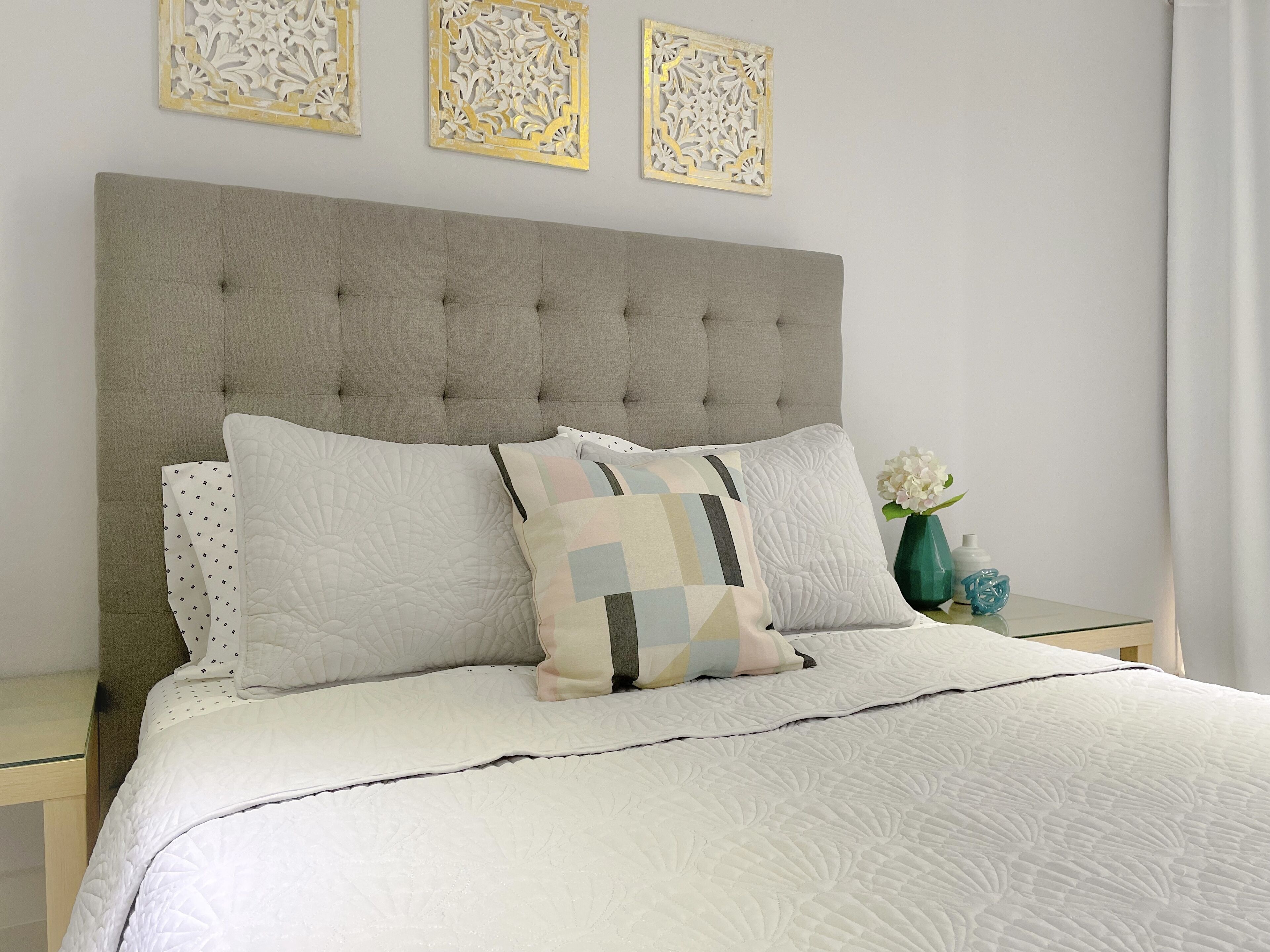 Un llit ben fet amb capçalera grisa tapissada, roba de llit blanca i coixins decoratius. Hi ha quadres a la pared sobre el llit i un ram de flors a la taula auxiliar.