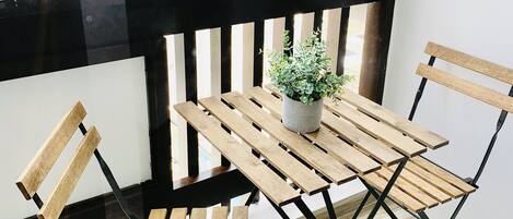 Un balcón con mesa y sillas de madera, una planta en maceta y vistas de un edificio con ventanas.