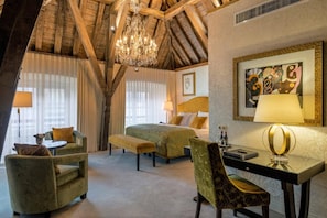 A hotel room with a wooden ceiling, a chandelier, a bed with green bedding, a desk with a lamp, and a painting on the wall.