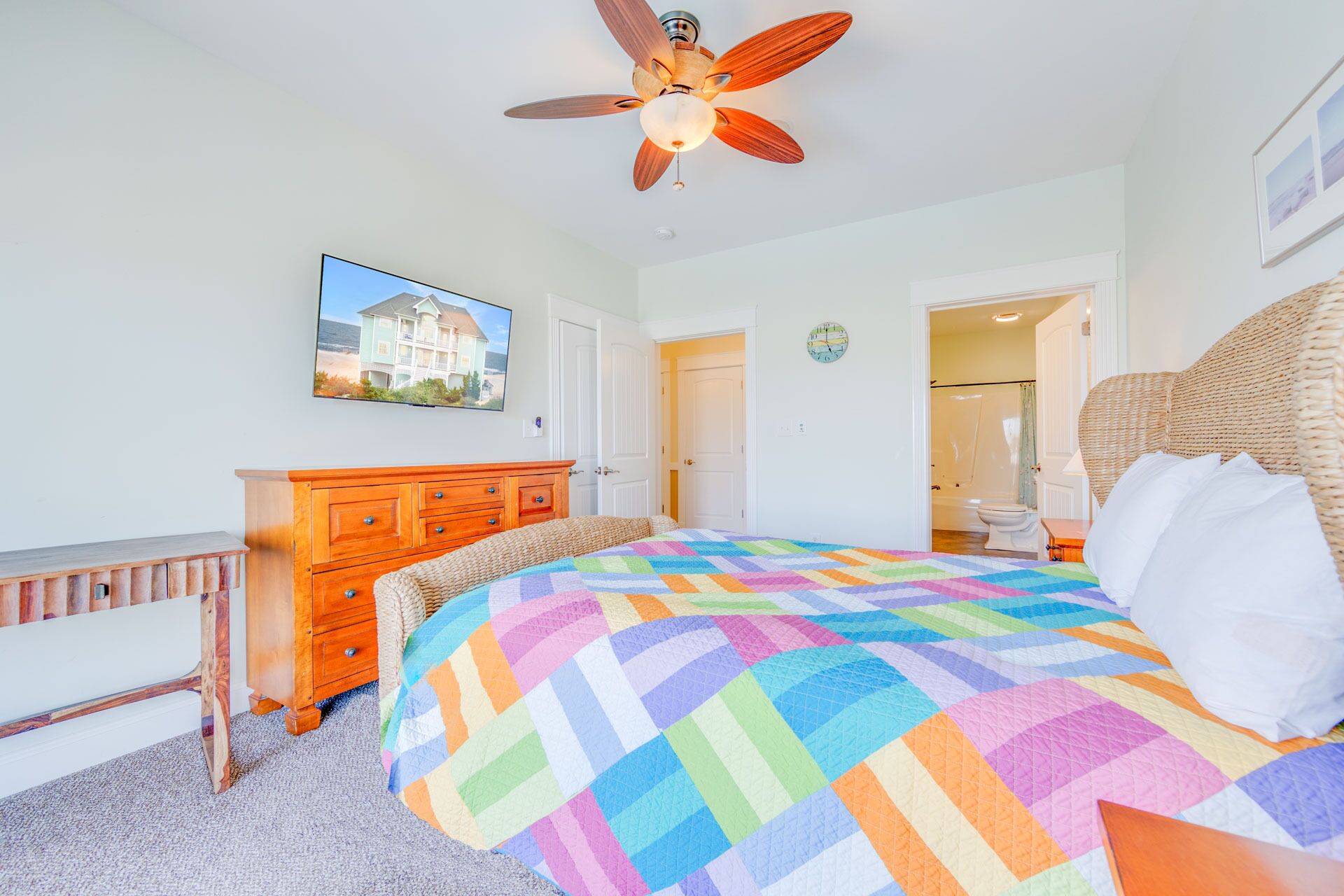 Een slaapkamer met een groot bed, een houten dressoir, een plafondventilator, een televisie en een badkamer die door een open deur zichtbaar is.