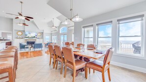 A spacious dining area with a large table and chairs, a kitchen in the background, and large windows offering a view of the ocean. - Premier Oceanfront Home w/ Pool, Elevator, Hot Tub, Theater Room (Avon)