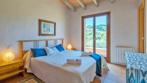 Un dormitorio con cama, lámparas de noche, un cuadro enmarco, una ventana con vistas y una mesa con mantel estampado.