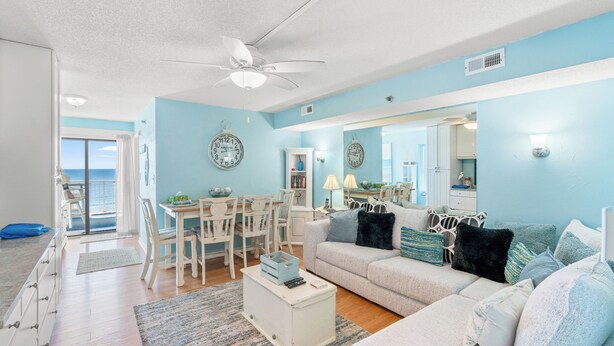 A living room with a white sofa, a coffee table, and a dining area with a white table and chairs. There is a ceiling fan and a view of the ocean through a sliding door. - Views & Indoor Pool - San A Bel 315 (North Myrtle Beach)
