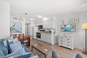 Een moderne woonkamer met een bank, salontafel en een eethoek. In de achtergrond is een keuken met witte kasten en apparatuur zichtbaar.