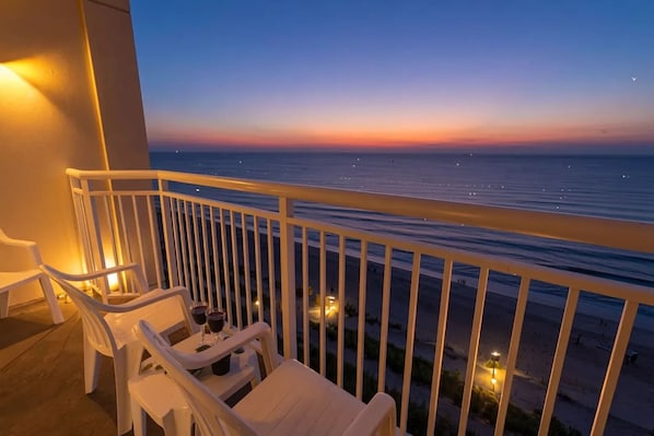 A balcony with white chairs and a table, overlooking a beach at sunset. - Holiday Sands South Beach 1007 - Ocean Front (Myrtle Beach)