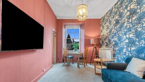 A living room with a large flat-screen TV, a blue sofa, a dining table with chairs, and a patterned wall.