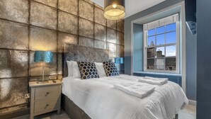 A neatly made bed with white linens and patterned pillows, a bedside lamp, and a view of a building through a window.