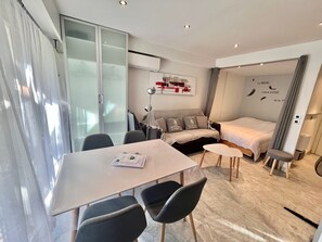 A modern hotel room with a glass door, a sofa bed, a small table, and a desk with chairs.