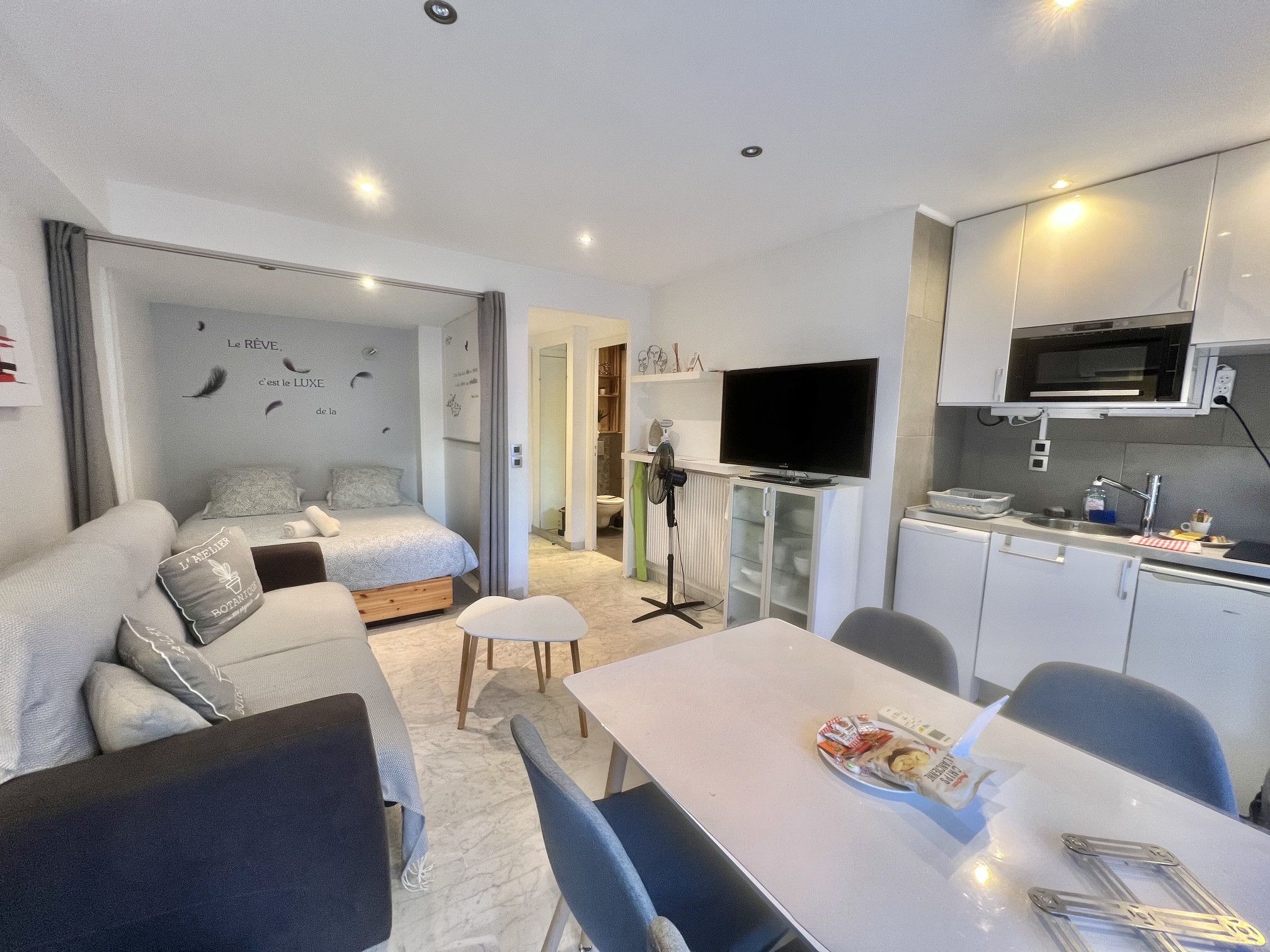 A compact hotel room with a bed, a dining table, a TV, and a kitchenette.